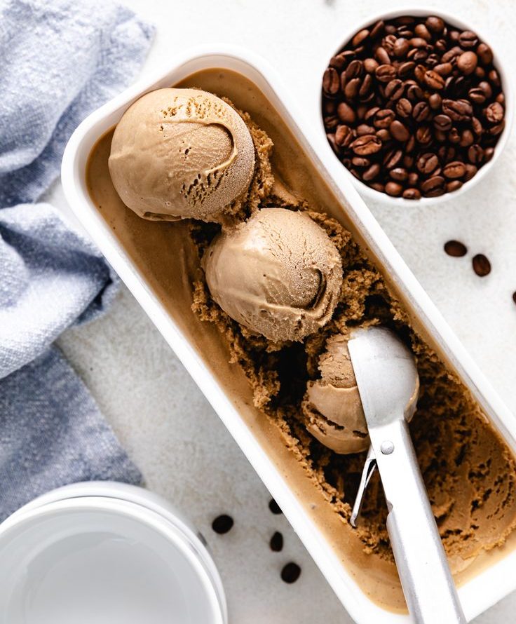 A tub of rich chocolate ice cream with three scoops and a metal scoop, surrounded by coffee beans and a white bowl.