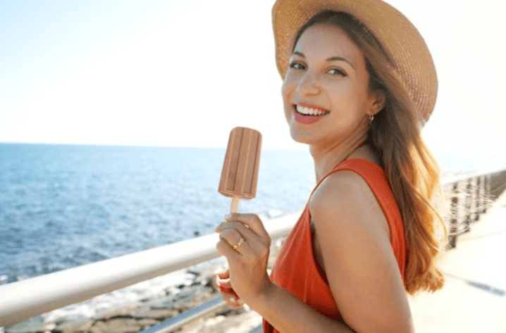 A person in a bright orange dress and a straw hat enjoys a popsicle, with a scenic ocean view in the background.